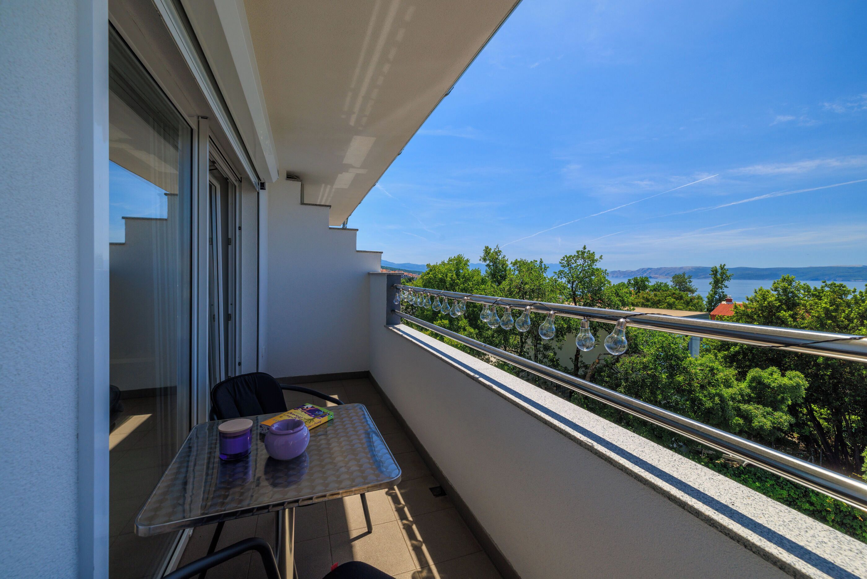 A balcony with a table, chairs, and a view of the sea and greenery.