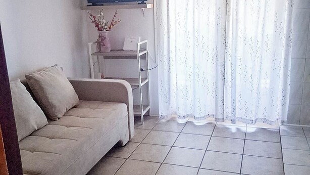 Ein Wohnzimmer mit einer Couch, einem an der Wand montierten Fernseher, einem Schreibtisch mit Stuhl und einem Fenster mit Vorhängen.