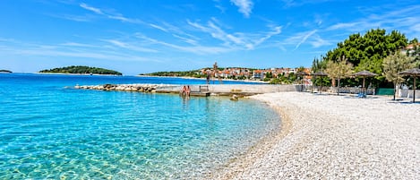A pebble beach with clear water, a pier, and a village in the background.