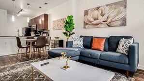 A modern living room with a blue sofa, a coffee table, and a dining area with chairs and a table. - Boulevard Stays Walk to Downtown! 2BR SoCo (Austin)