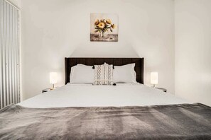 A neatly made bed with a dark headboard, two bedside lamps, and a framed artwork of flowers above it.