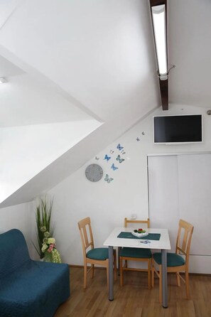 Ein Zimmer mit schrägem Dach, einem an der Wand befestigten Fernseher, einem Esstisch mit Stühlen und einem blauen Sofa.
