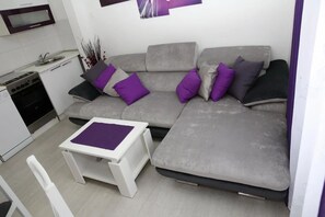 Ein kompakter Wohnbereich mit grauem Sofa, einem kleinen weißen Tisch und einer Küchenzeile mit Herd und Spüle.