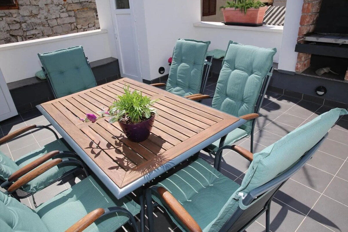 Eine Außenpatio mit einem Holztisch, grünen Sitzgelegenheiten mit Polstern, einem Topfpflanzen und einem Backofen aus Ziegeln.