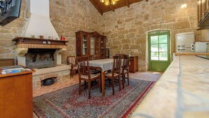 Um quarto com paredes de pedra, uma lareira, uma mesa de jantar com cadeiras, uma porta verde e uma área de cozinha.
