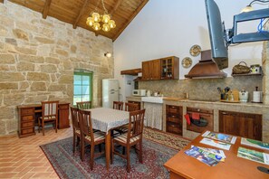 A rustic kitchen with stone walls, wooden cabinets, a dining table with chairs, and a hanging light fixture. - House Barba Frane (Vizinada)