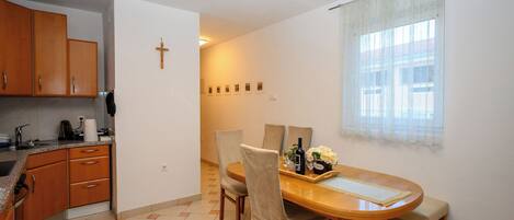 A dining area with a wooden table, chairs, and a bench. There is a kitchen area with wooden cabinets and a cross on the wall.