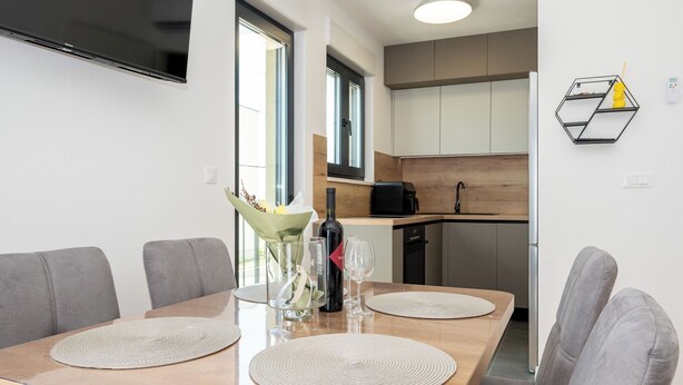 Un espace repas moderne avec une table en bois, des chaises à revêtement gris, un téléviseur fixé au mur et une cuisine en arrière-plan.