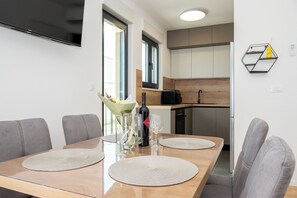 A modern dining area with a wooden table, grey upholstered chairs, a TV mounted on the wall, and a kitchen in the background. - Apartment Seaview Terrace (Podstrana)