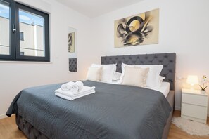 A neatly made bed with a grey quilt, white pillows, and rolled towels on a white nightstand with a lamp and a small potted plant. - Apartment Seaview Terrace (Podstrana)