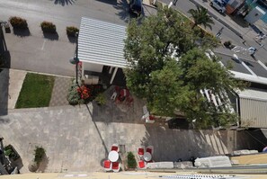 An aerial view of a building with a flat roof, outdoor seating area with red chairs and tables, and a large tree in front. - Apartment Nikola Centar (Biograd na Moru)