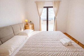 A bedroom with a bed, a nightstand, a lamp, and a view through a window. - Apartment Franky (Crikvenica)
