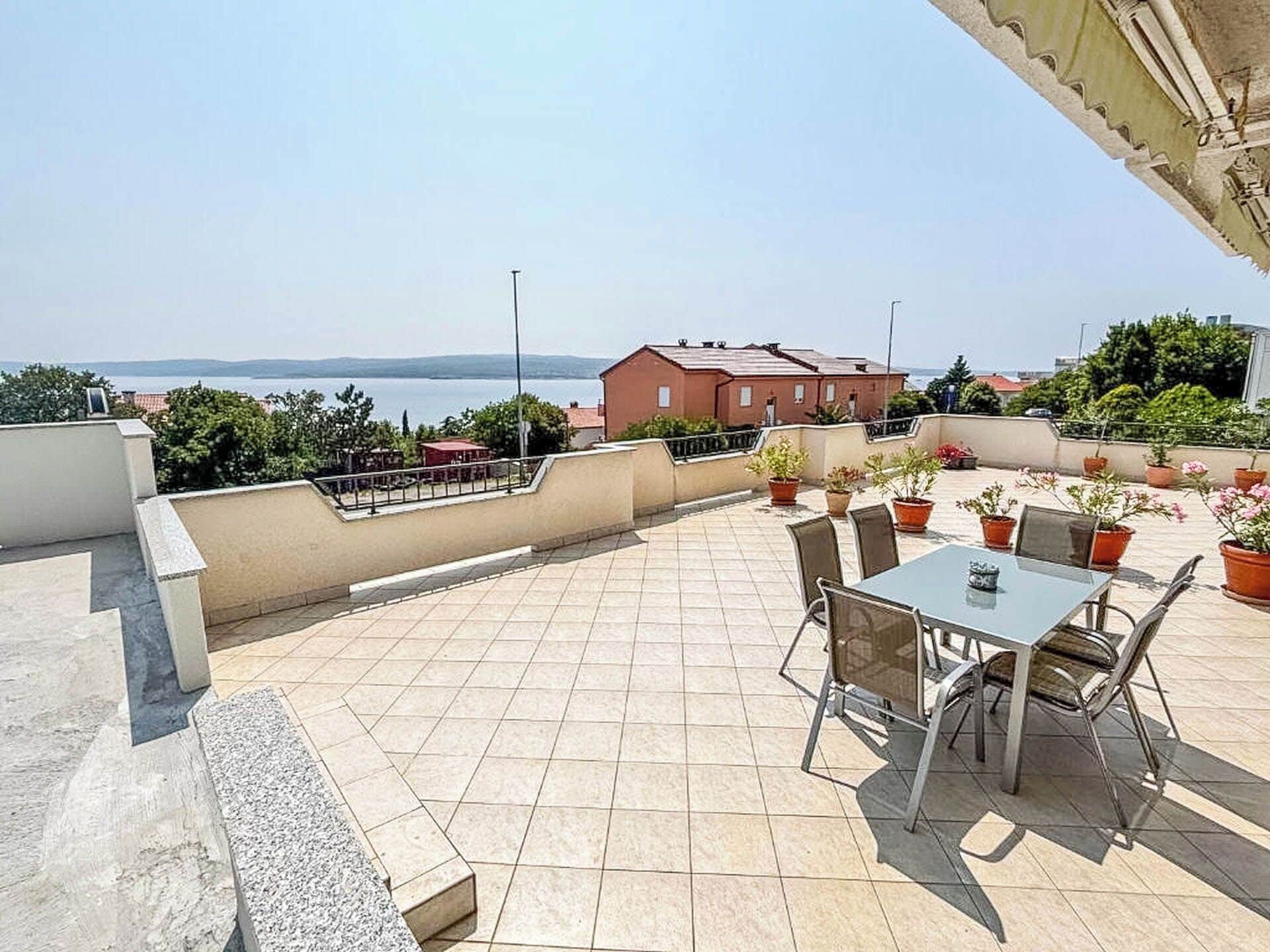 A rooftop terrace with a table and chairs, potted plants, and a view of the sea.