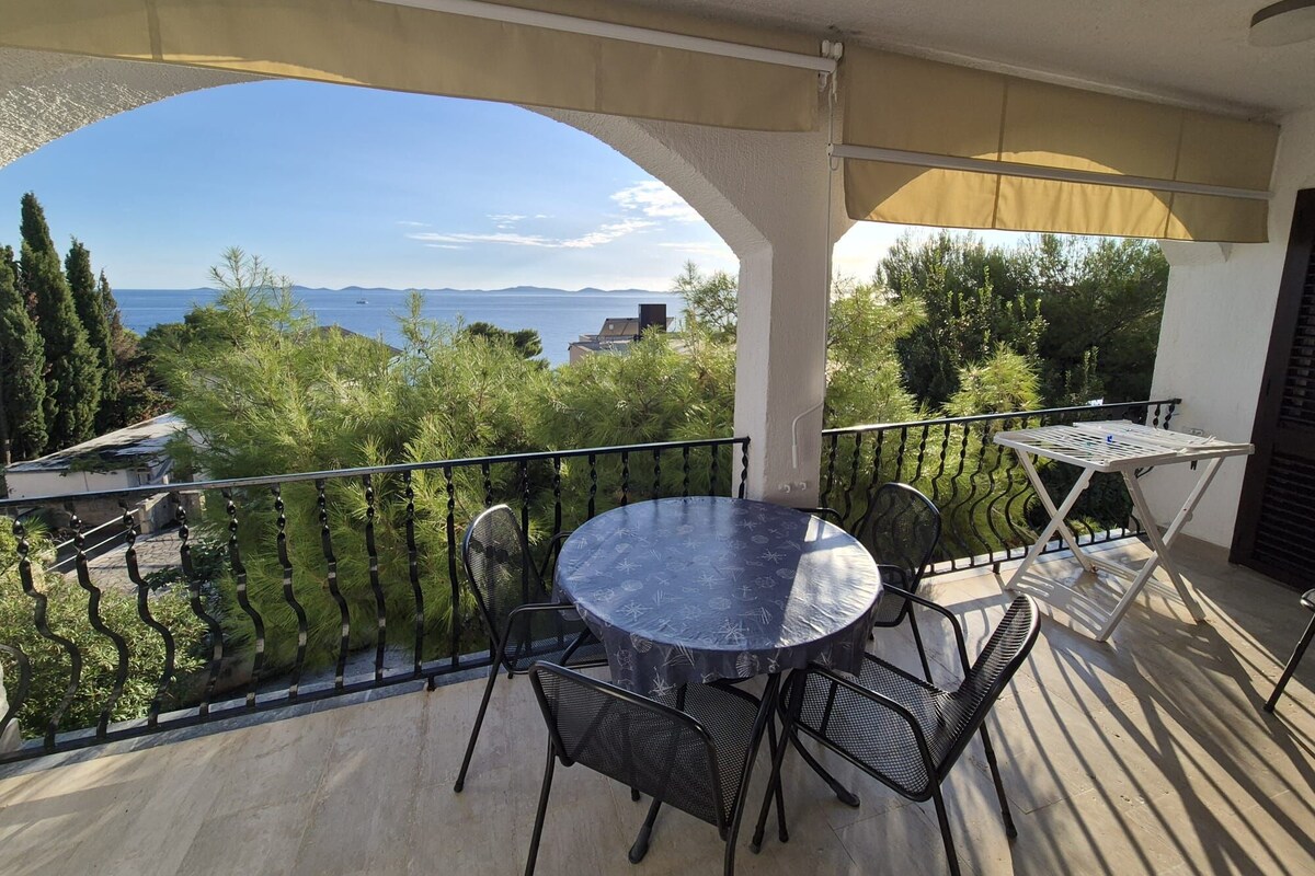 A balcony with a round table and chairs, a view of the sea, and a covered area with a pergola.