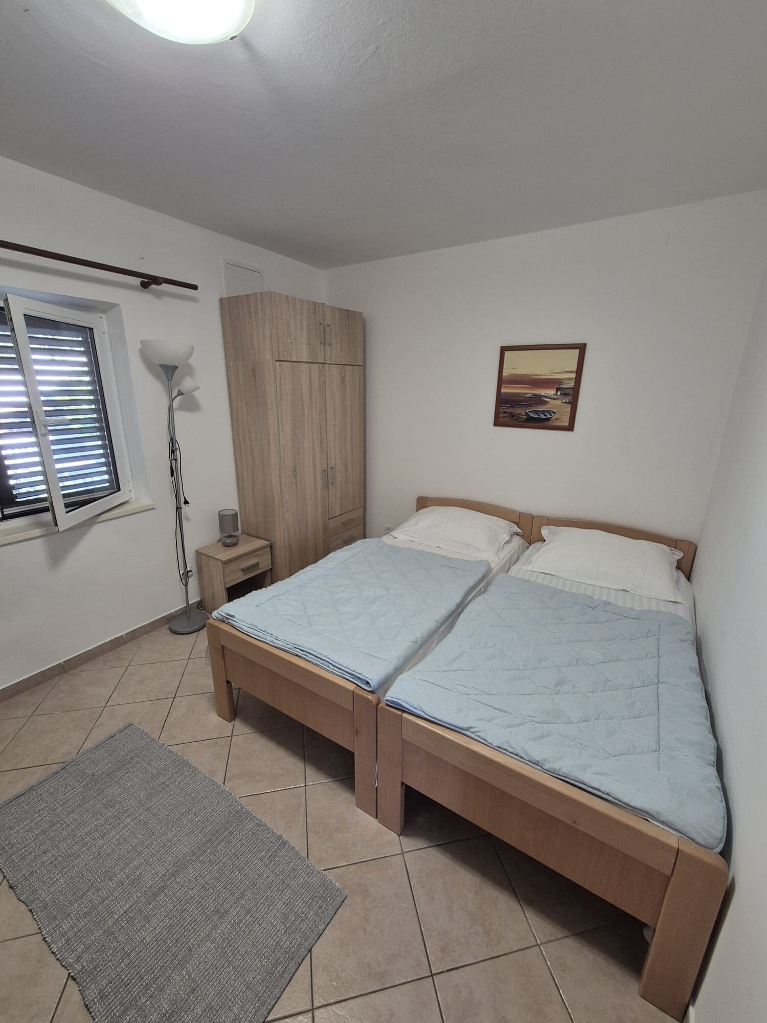 A bedroom with a bed, a wardrobe, a bedside table, a lamp, and a window with blinds.