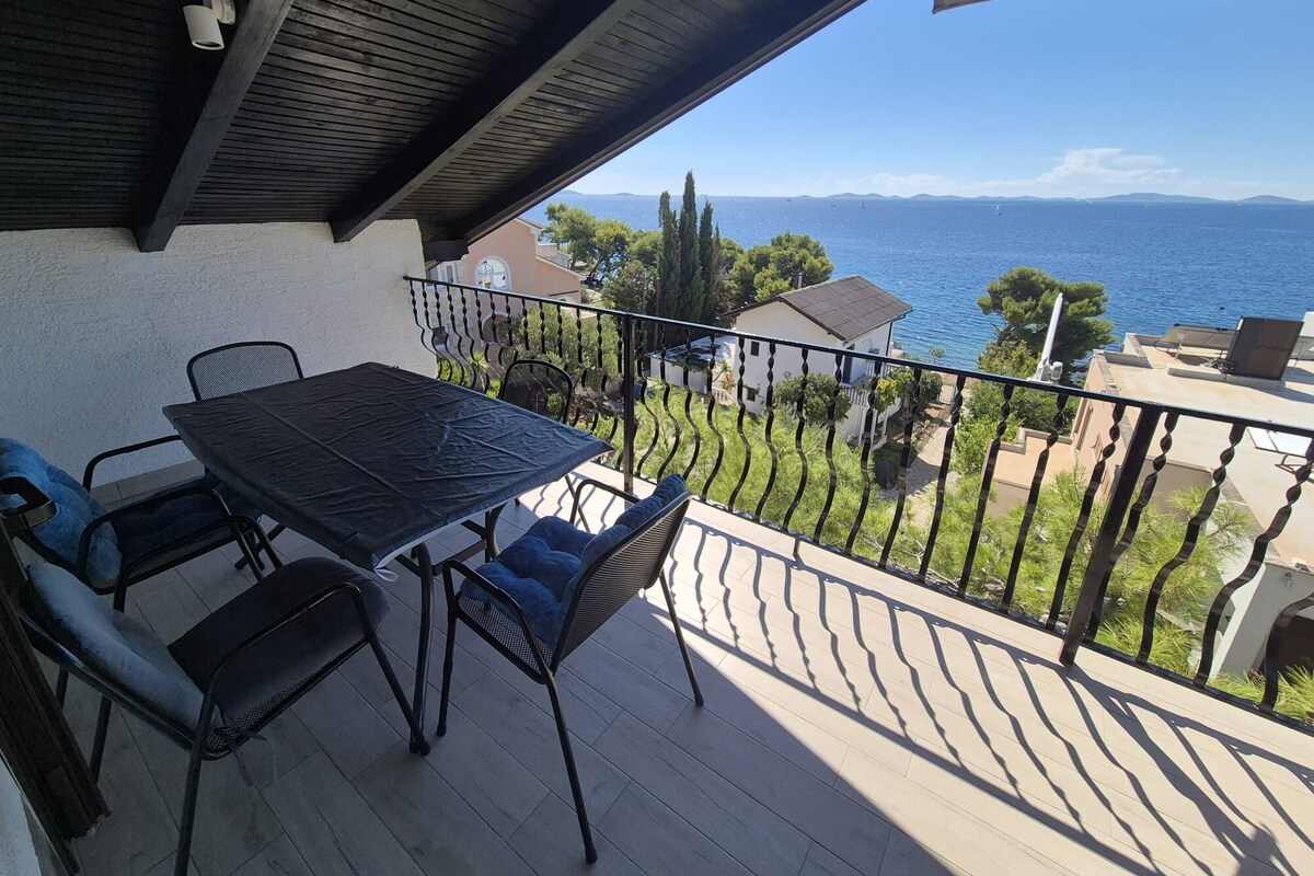 A balcony with a table and chairs, overlooking a body of water and a distant coastline.