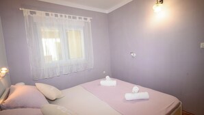 A bedroom with a bed, pillows, a window with a sheer curtain, a wall-mounted light, and a bedside table. - Apartment Nikol (Crikvenica)