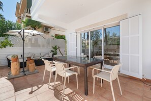 En udendørs terrasse med et glasbord, hvide stole og en parasol.