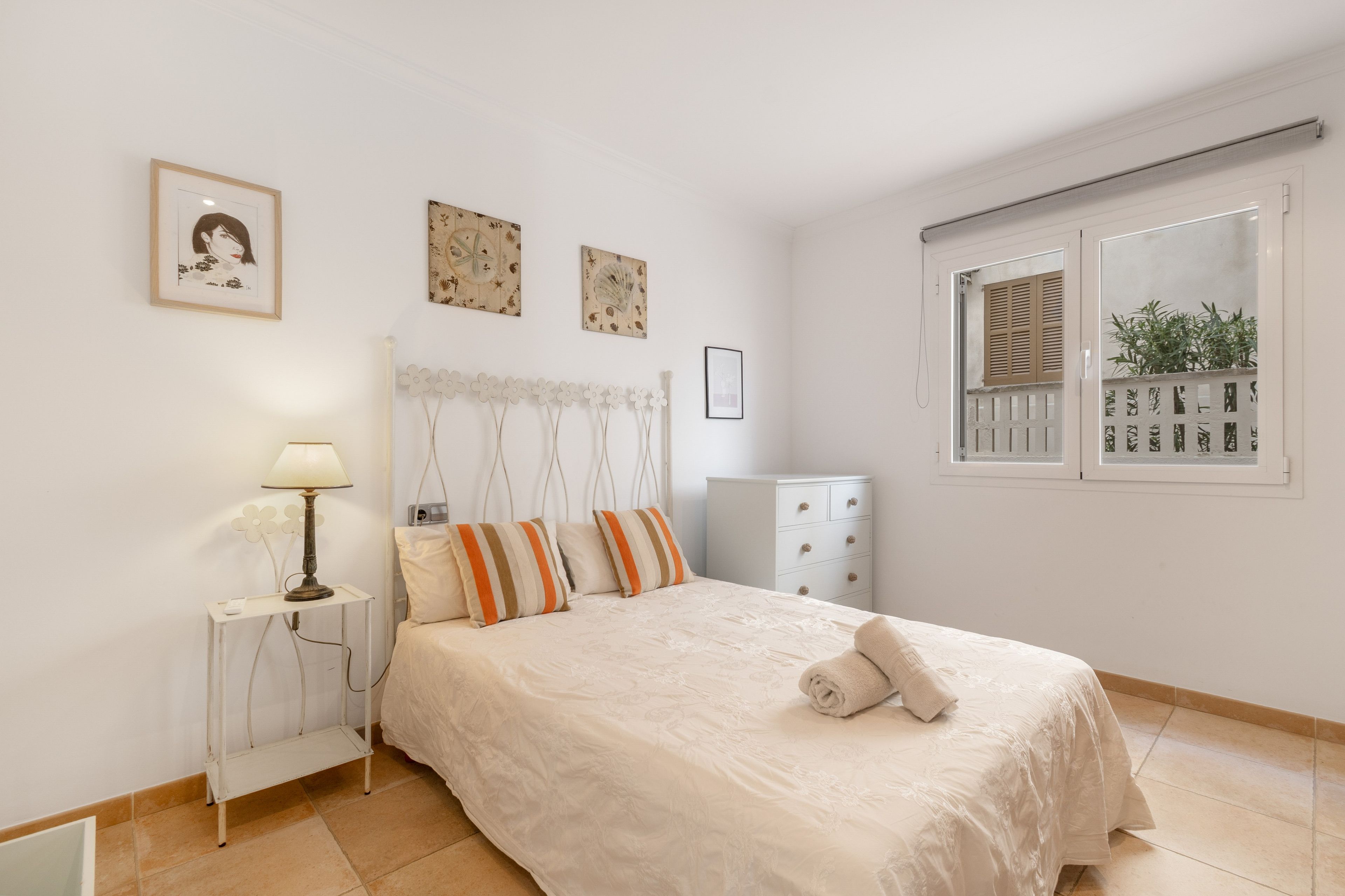 A bedroom with a bed, a nightstand, a lamp, a dresser, and a window with shutters.