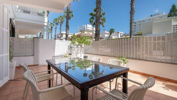 A balcony with a glass table and chairs, surrounded by a white fence, palm trees, and buildings in the background.
