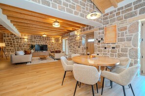 Un vaste espace de vie agrémenté d’un mur en pierre, de poutres apparentes en bois au plafond, d’une table de salle à manger ronde en bois et d’un téléviseur à écran plat.
