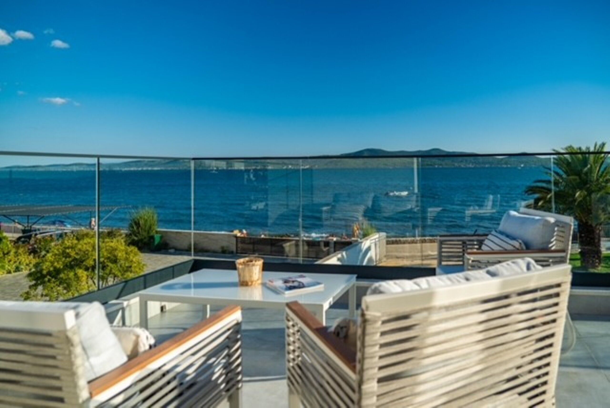 A terrace with outdoor furniture, a glass balustrade, and a view of the sea and distant land.