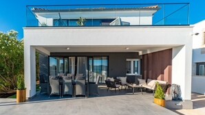 Een modern huis met twee verdiepingen, voorzien van een overdekte patio met buitenmeubilair en een glazen balustrade.
