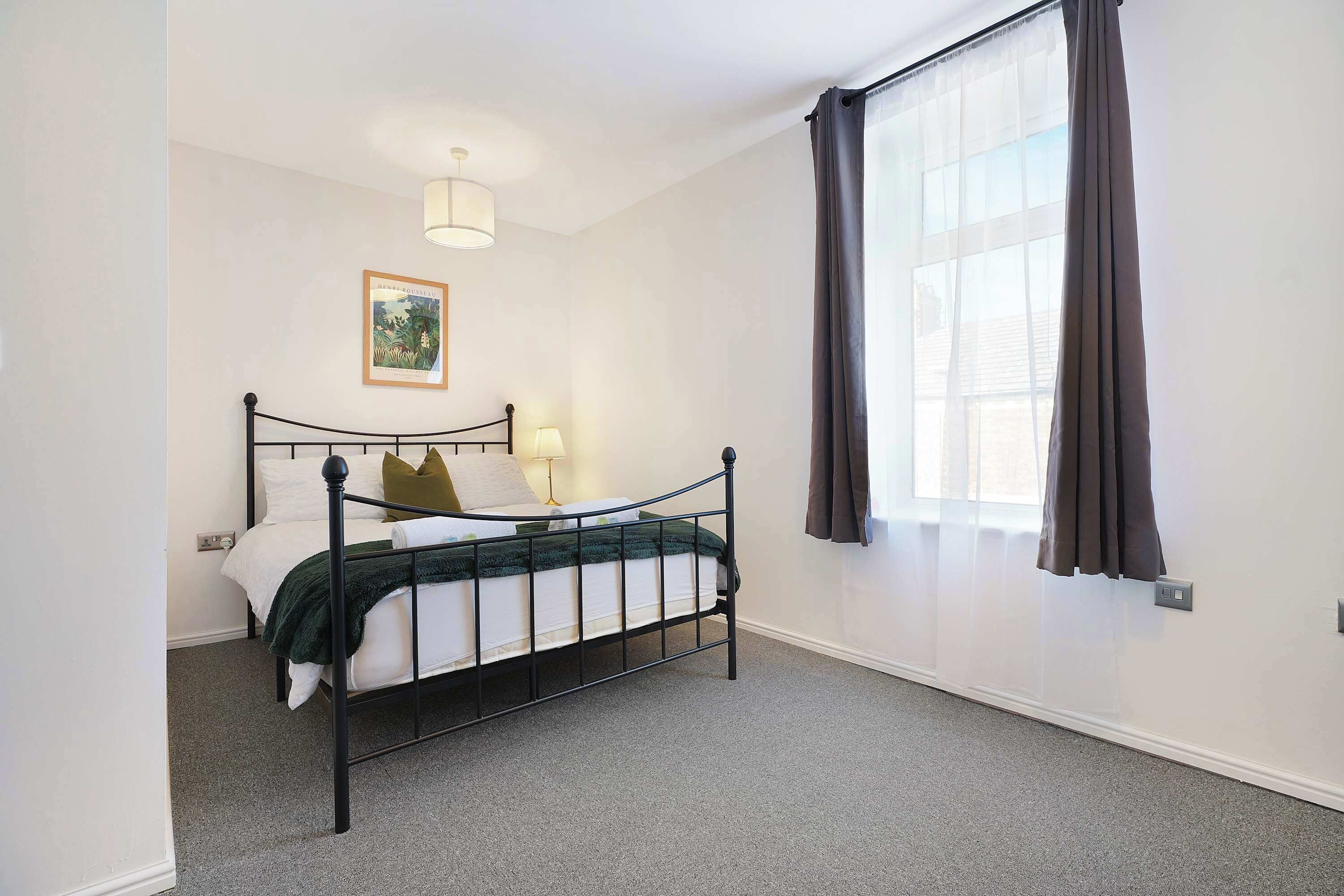 A bedroom with a bed, a window with curtains, a wall-mounted light, and a framed picture.