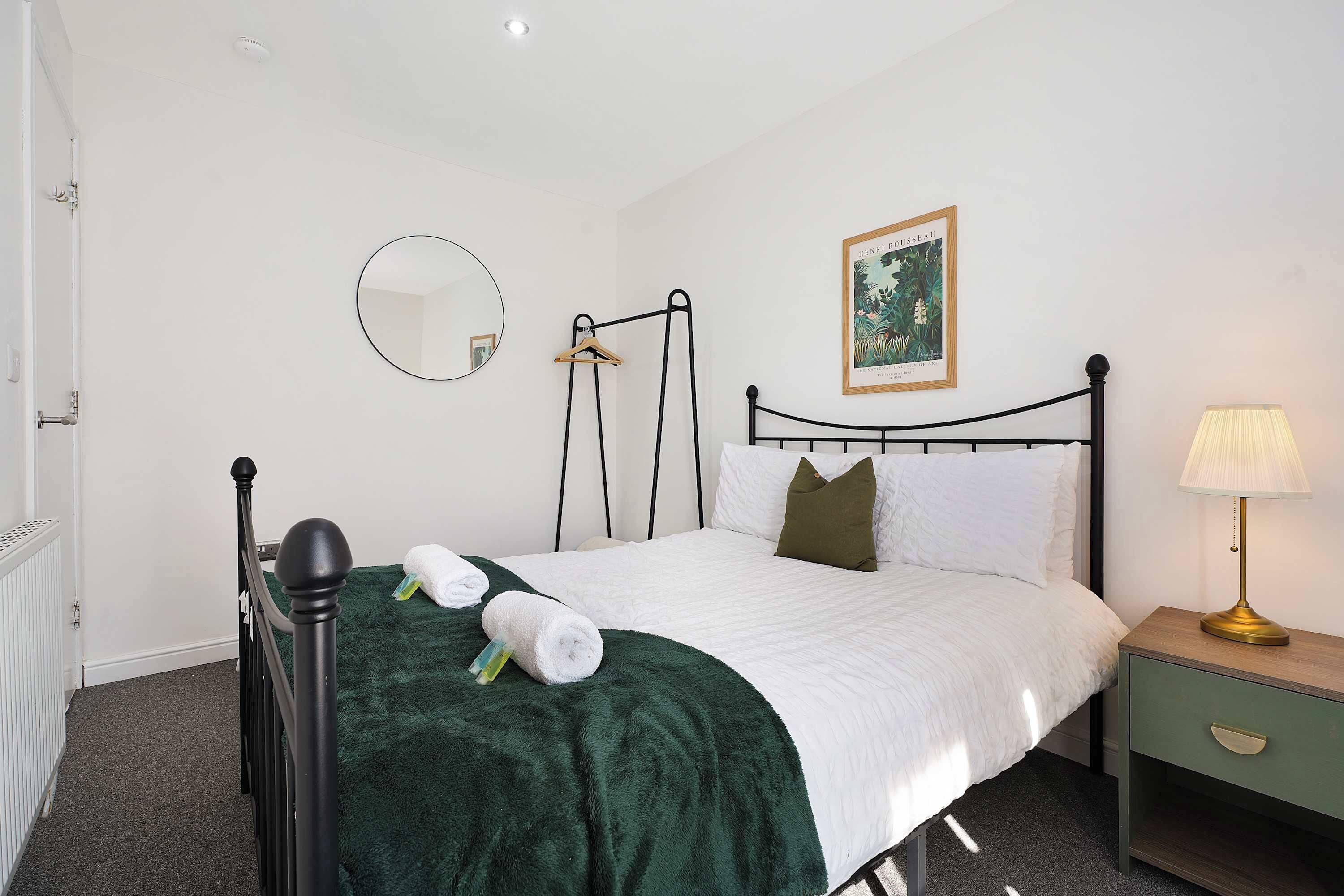 A neatly made bed with a green blanket, a bedside table with a lamp, a hanging coat rack, a framed picture on the wall, and a round mirror.