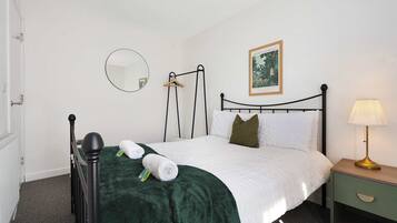 A neatly made bed with a green blanket, a bedside table with a lamp, a hanging coat rack, a framed picture on the wall, and a round mirror.