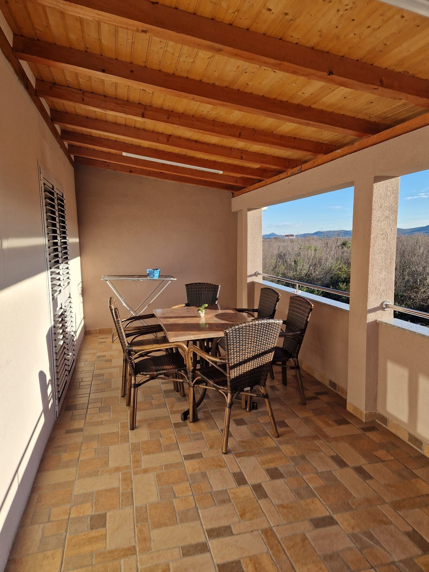 A covered patio with a table and chairs, a view of the landscape, and a window with blinds.