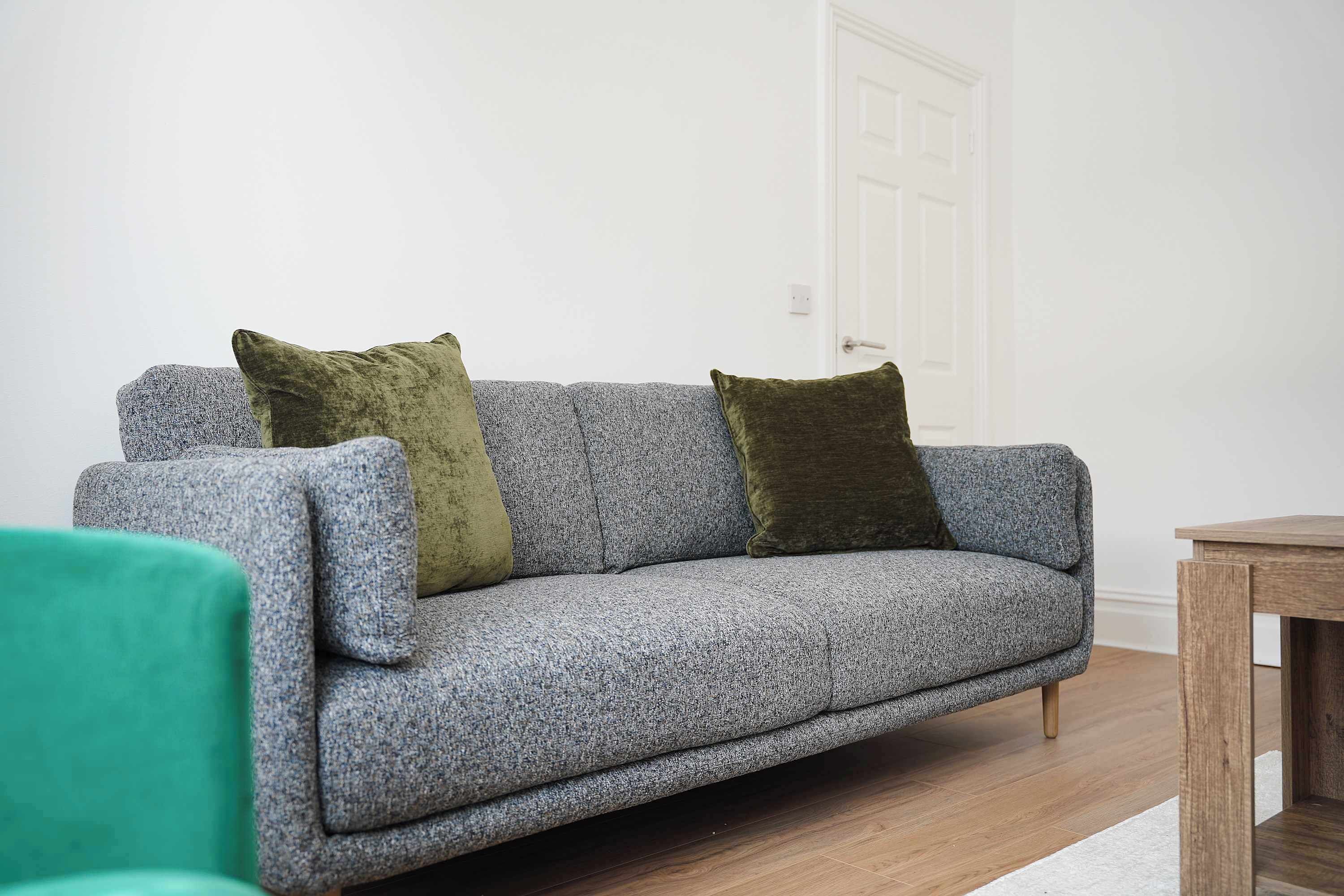 A grey sofa with two cushions, a wooden side table, and a white door in a room with wooden flooring.