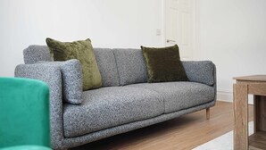 Un sofá gris con dos cojines, una mesita de madera y una puerta blanca en un cuarto con piso de madera.