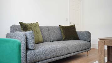 A grey sofa with two cushions, a wooden side table, and a white door in a room with wooden flooring.
