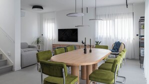 A modern dining area with a round table, green upholstered chairs, pendant lights, and a large flat-screen TV on the wall. - Villa Respiro Residence South (Kastela)
