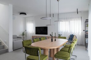 A modern dining area with a round table, green upholstered chairs, pendant lights, and a large flat-screen TV on the wall. - Villa Respiro Residence South (Kastela)