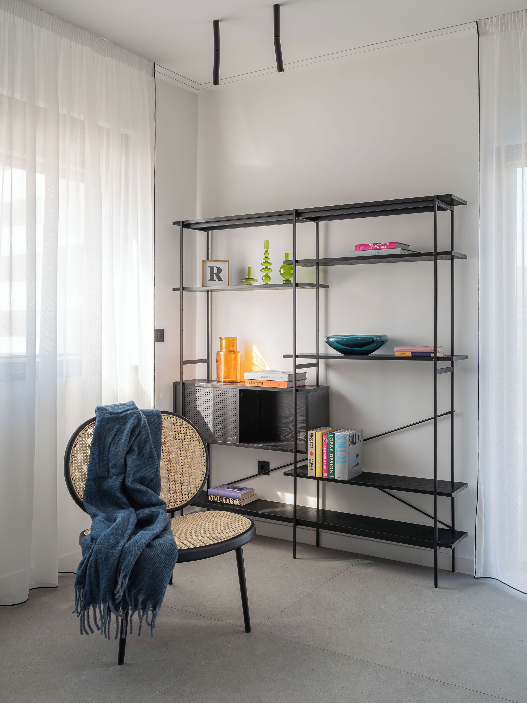 Una habitación moderna con una estantería de metal negro, un sillón de mimbre con una manta azul y un ventanal con cortinas de gasa.