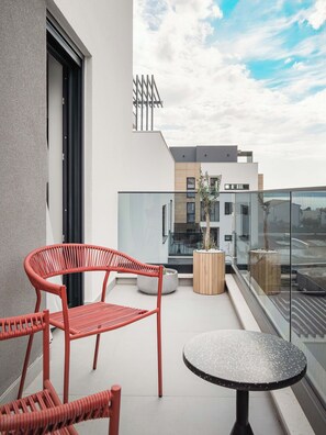 Un balcon doté d’une balustrade en verre, d’une table ronde et de deux chaises en osier rouge.