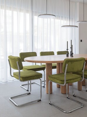 Un espace repas comprenant une table ronde, six chaises à l’assise rembourrée de tissu vert et deux lampes suspendues.