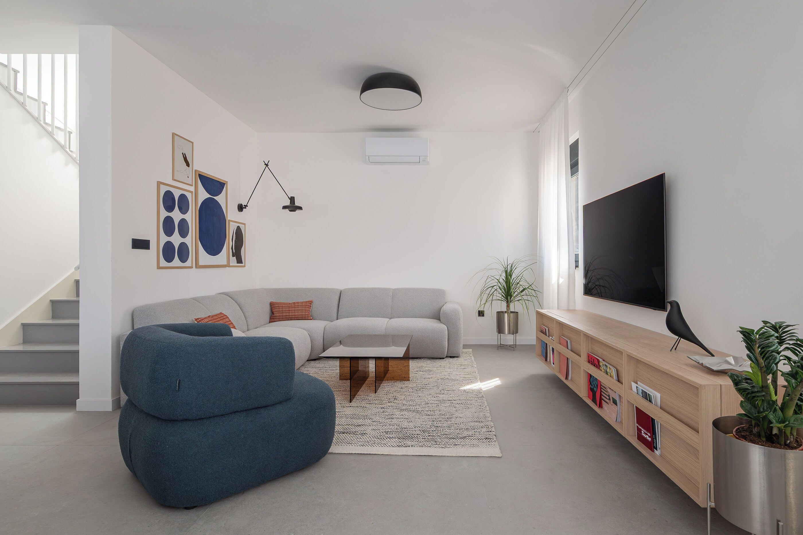 Ein modernes Wohnzimmer mit einem grauen Ecksofa, einem blauen Sessel, einem Holztisch, einem Flachbildfernseher, einem Bücherregal und einer Treppe.