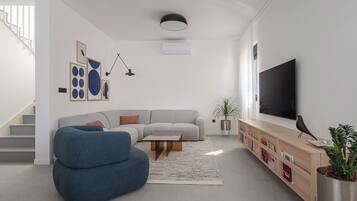 Una sala moderna con un sofá seccional gris, un sillón azul, una mesa de centro de madera, un televisor de pantalla plana, una estantería y una escalera.
