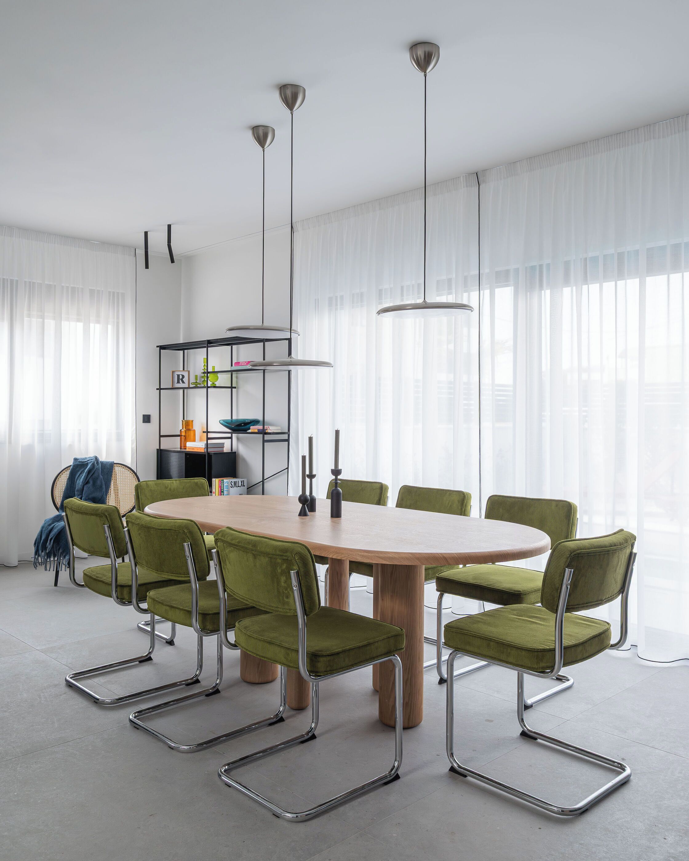 Ein zeitgemäßes Esszimmer mit einem Holztisch, mit dunkelgrünen Stoffstühlen, einem schwarzen Metallregal und Pendelleuchten.