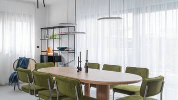 A modern dining room with a wooden table and green upholstered chairs, a black metal shelving unit, and pendant lighting.