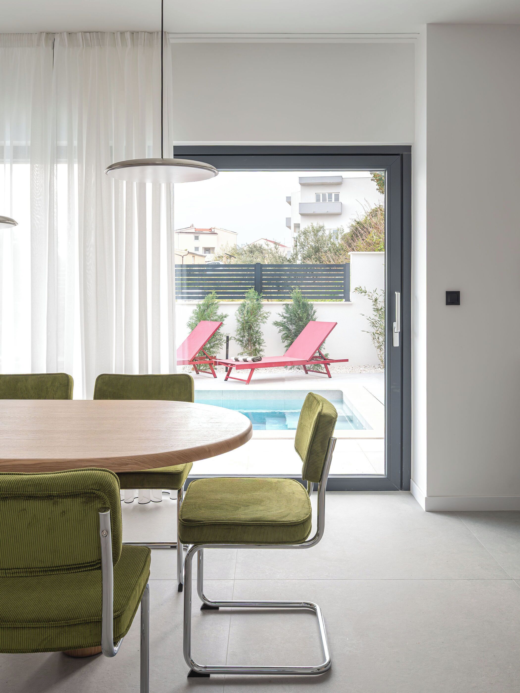 A modern room with a round wooden table, green upholstered chairs, a glass door leading to an outdoor area with a pool and red lounge chairs.