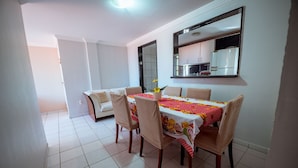 A dining area with a table set for four, surrounded by chairs, in a room with a tiled floor and a kitchen visible through an open doorway. - Apto c 3 Quartos e Garagem a 260 m da Praia (João Pessoa)