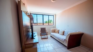 A living room with a sofa, a chair, a TV, and a window with a view of palm trees. - Apto c 3 Quartos e Garagem a 260 m da Praia (João Pessoa)