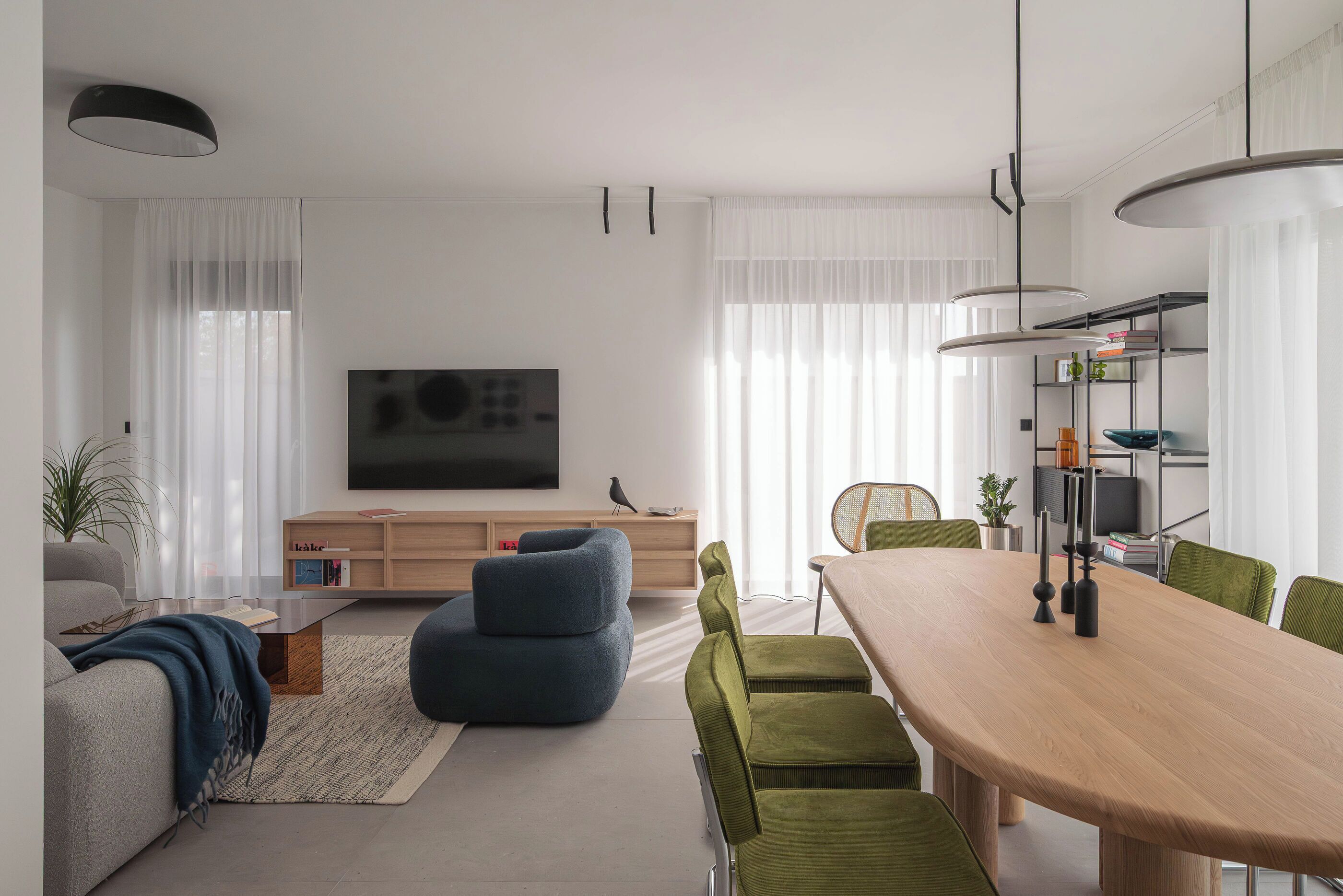 A modern living room with a flat-screen TV, a wooden dining table, green chairs, and a blue sofa.
