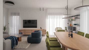 A modern living room with a flat-screen TV, a wooden dining table, green chairs, and a blue sofa.
