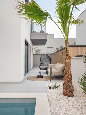 A modern outdoor patio with a pool, a palm tree, a round table, and a chair. - Villa Respiro Residence North (Kastela)