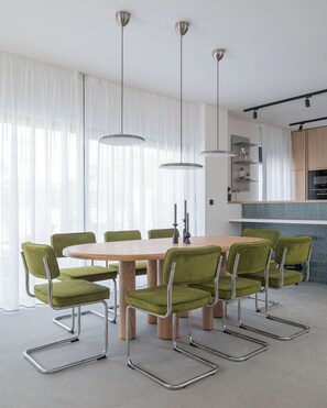 A modern dining area with a round table, green upholstered chairs, pendant lighting, and a kitchen in the background. - Villa Respiro Residence North (Kastela)